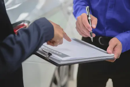 Duas pessoas de pé, uma segurando uma prancheta e caneta na mão e a outra apontando para o papel