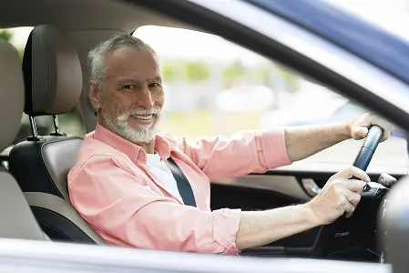 Homem de cabelo grisalho, com camisa rosa e cinto de segurança, sentado ao volante e sorrindo