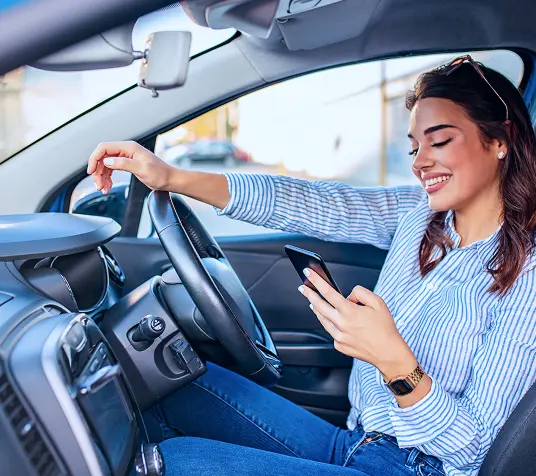 A imagem mostra o interior de um carro. Nele, há uma mulher branca, sorrindo enquanto segura um celular.