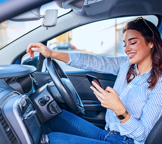 A imagem mostra uma mulher de pele branca sorrindo e olhando pela janela do seu carro, no banco do motorista. Com a mão direita ela segura o volante e com a mão esquerda está segurando e mostrando a chave do carro. Ela tem o cabelo escuro, curto e encaracolado, está usando um brinco discreto de formas geométricas em tons azuis e uma blusa na cor rosa salmão.