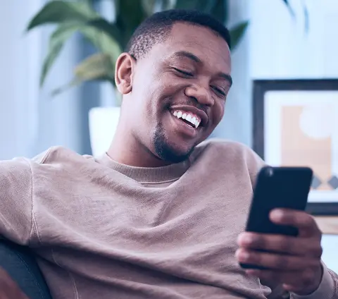 A imagem tem um homem negro sorrindo enquanto olha para o celular. Ele está em uma sala, sentado em um sofá.