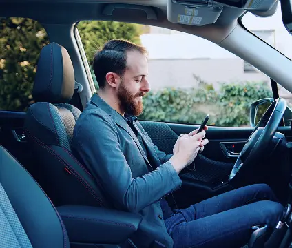 A imagem mostra o interior de um carro. Nele, há um homem branco, sorrindo enquanto segura um celular.