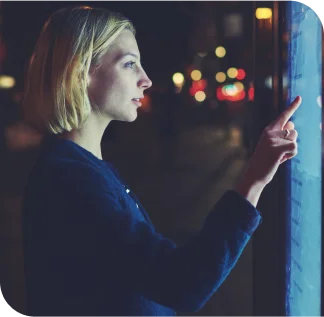 Em uma rua, à noite, há uma tela touchscreen de lado e uma mulher loira de cabelo curto e blusa azul marinho interage com ela com seu dedo indicador.
