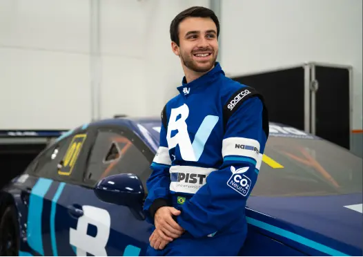 O piloto Felipe Baptista está encostado no carro de corrida, ele sorri e veste um macacão azul com o logo do banco BV.