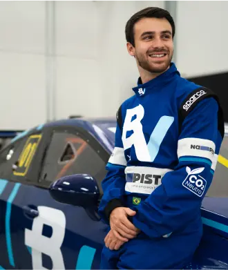 O piloto Felipe Baptista está encostado no carro de corrida, ele sorri e veste um macacão azul com o logo do banco BV.