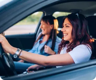 Duas mulheres alegres dentro de um carro, uma delas está conduzindo e a outra no banco do passageiro.