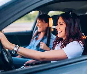 Duas mulheres alegres dentro de um carro, uma delas está conduzindo e a outra no banco do passageiro.