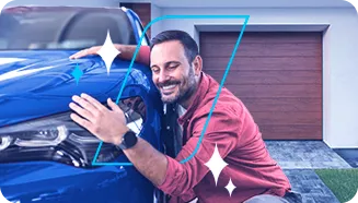 Homem feliz agachado abraçando um automóvel azul em frente uma garagem.