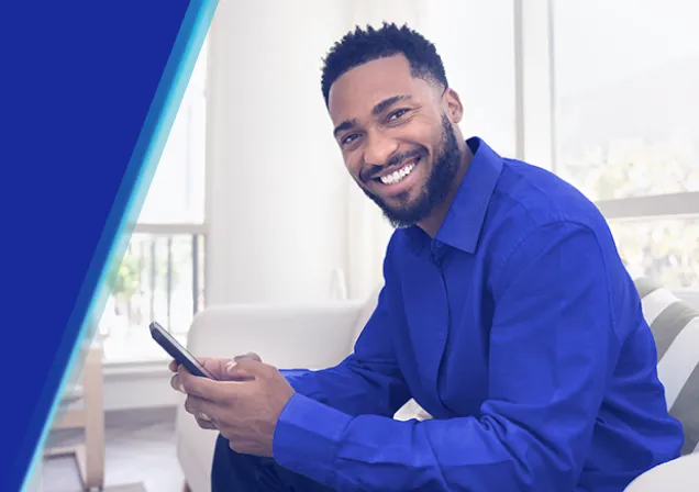 Homem sorridente, com camisa azul, segurando um celular. Ele está sentado em um ambiente iluminado.