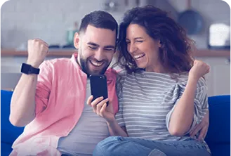 Na imagem, temos um homem e uma mulher sentados no sofá felizes, ambos olhando para o celular que a mulher está segurando.