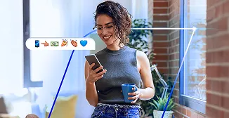 Mulher sorrindo em um escritório, usa o celular e segura uma caneca. Uma faixa de emojis sai do aparelho.