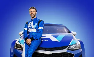 Um piloto sorrindo, com macacão do banco BV. Ele está apoiado em um carro de corrida com fundo azul.