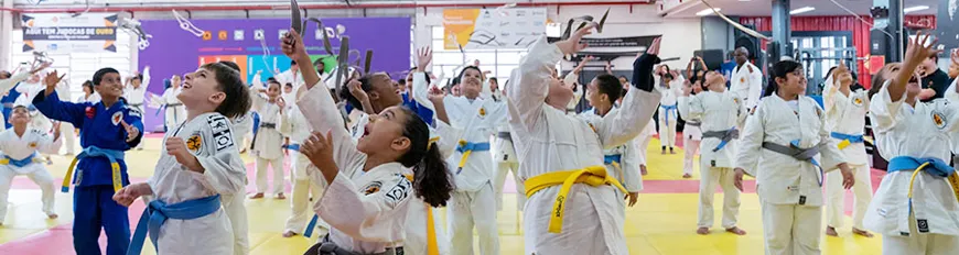 Várias crianças com quimonos e faixas de jiu-jitsu, em um tatame, comemorando felizes.