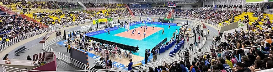 Uma quadra de vôlei, com atletas jogando, e várias pessoas na arquibancada torcendo e assistindo.