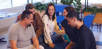 Grupo de cinco colegas conversando e sorrindo em área informal de escritório, tom descontraído e colaborativo.