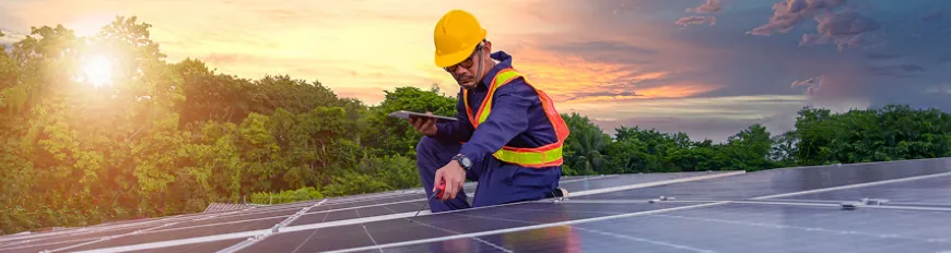 Técnico com capacete e colete inspeciona painel solar ao pôr do sol, ele está segurando um tablet e ferramenta.
