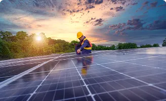 Técnico com capacete e colete inspeciona painel solar ao pôr do sol, ele está segurando um tablet e ferramenta.