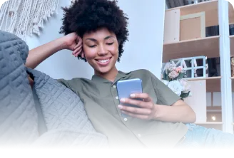 Mulher sorrindo recostada no sofá, usando camisa verde, segurando um celular em ambiente interno com estante ao fundo.