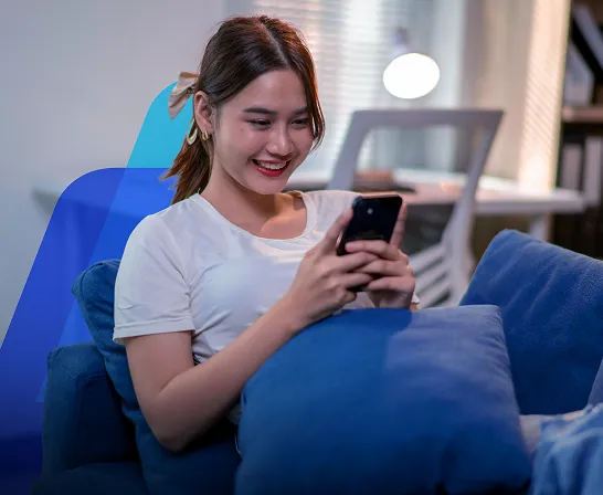Fotografia de uma mulher jovem de cabelo escuro preso, sentada em um sofá azul em uma sala com uma luminária acesa ao fundo. Ela sorri enquanto mexe no celular.