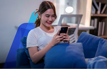 Fotografia de uma mulher jovem de cabelo escuro preso, sentada em um sofá azul em uma sala com uma luminária acesa ao fundo. Ela sorri enquanto mexe no celular.