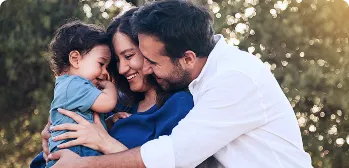 Um casal de um homem e uma mulher sorrindo abraçam um bebê em um fundo com árvores.