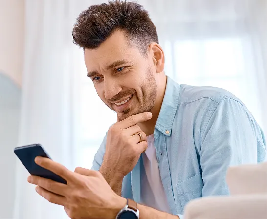 Um homem branco, de cabelos curtos, segura o celular com umas das mãos e sorri. 