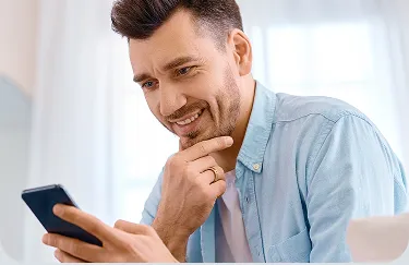 Um homem branco, de cabelos curtos, segura o celular com umas das mãos e sorri. 