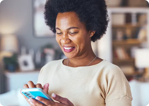 Uma mulher negra de cabelos cacheados curtos, segura o celular com as duas mãos e sorri. 