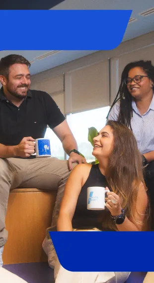 Um homem e duas mulheres sentados, sorrindo de maneira descontraída e segurando uma caneca com a logo do banco BV.