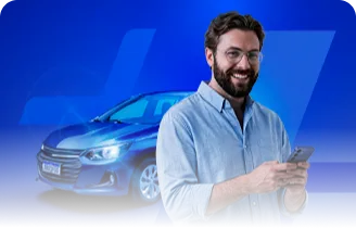 Homem branco em pé, vestindo uma camisa de botão azul-clara, com o celular na mão. Logo atrás dele, ao fundo, um carro azul.