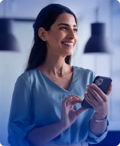 Mulher branca, em pé, vestindo uma camisa azul, com o celular na mão e sorrindo.