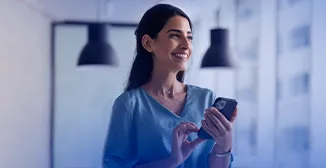 Mulher branca, em pé, vestindo uma camisa azul, com o celular na mão e sorrindo.