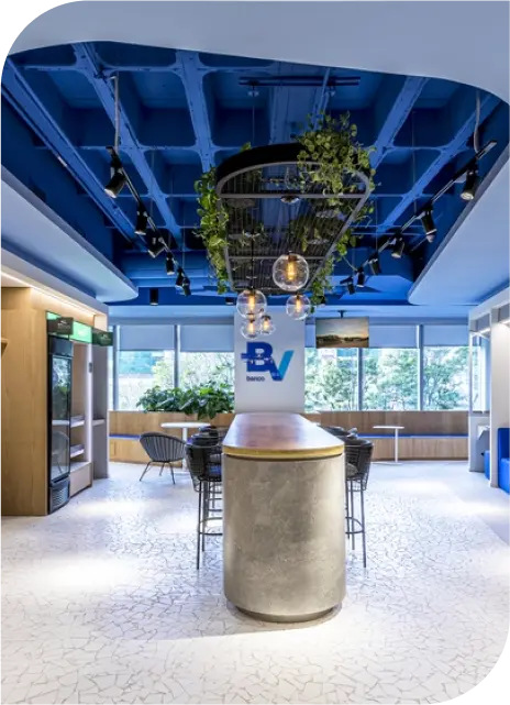 Copa do escritório BV com balcão e banquetas, logo do banco acima, mesas à direita e lustre central com plantas decorativas