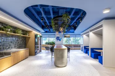 Copa do escritório do banco BV, onde apresenta um balcão central grande com algumas banquetas com o logo do banco logo acima, algumas mesas separadas a direita, e uma pia grande á esquerda com uma decoração com plantas.