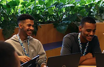 Ao fundo uma floreira em madeira abriga folhas verdes. Dois homens negros estão sentados sorrindo em frente a um notebook.
