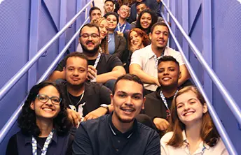 Em uma escada, com paredes azuis aos lados, 15 pessoas sorridentes estão sentadas nos degraus.