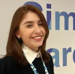Uma mulher banca de cabelos pretos e longos com franja, usa uma blusa cinza de gola alta e sorri.