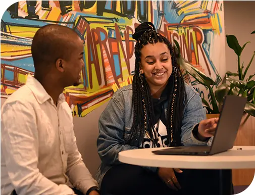Homem e mulher sorridentes em frente a uma mesa branca com um notebook aberto enquanto a mulher aponta para o notebook.
