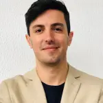 Um homem branco de cabelos curtos usa um terno bege, camiseta preta e esboça um sorriso.