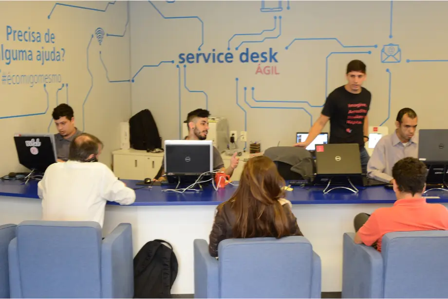 Na área de service desk, três pessoas estão sentadas de costas em poltronas azuis. Três pessoas estão sentadas em frente a uma bancada azul, trabalhando em computadores. Uma pessoa está em pé com a mão na cadeira.