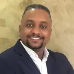 Um homem negro tem os cabelos curtos, um cavanhaque com bigode, usa um terno preto com camisa branca e sorri.