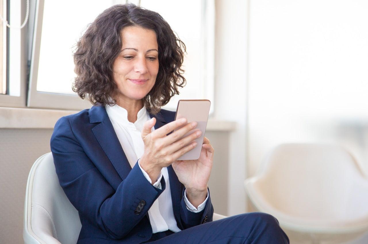 Mulher com semblante feliz pesquisando sobre Antecipação de Recebíveis em seu celular. Ela está sentada em uma cadeira branca e usando um blazer azul marinho.