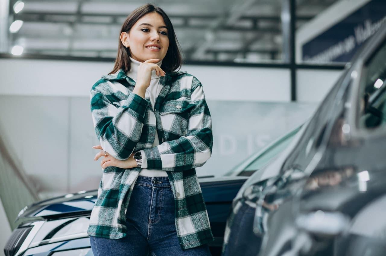 Mulher sorridente, com as mãos apoiadas no queixo, observando veículo para comprar direto com o proprietário. Ela veste sobretudo xadrez e calça jeans.