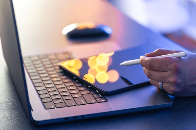 Mão segurando uma caneta stylus sobre uma tablet colocada sobre um teclado de laptop, com reflexos de luzes na tela e mouse ao fundo — ambiente de trabalho digital.
