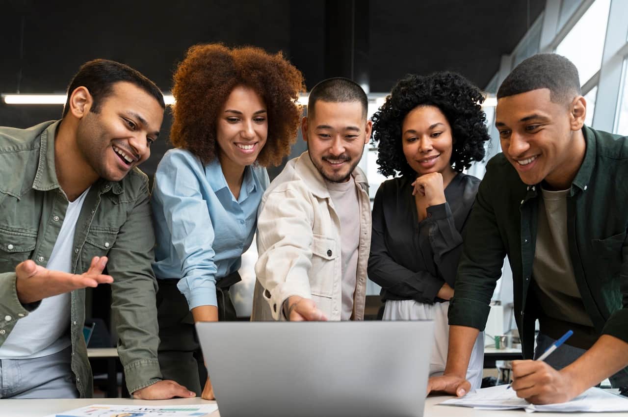 Grupo diverso de cinco jovens profissionais sorrindo e olhando para a tela de um notebook que está sobre uma mesa. Eles representam o capital humano da empresa onde trabalham.