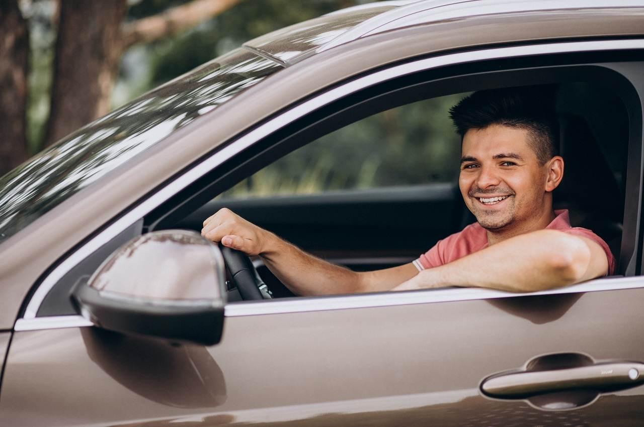 Homem dirigindo um carro, sorrindo, com clima de outdoor e uma expressão feliz ao volante em um ambiente ao ar livre.