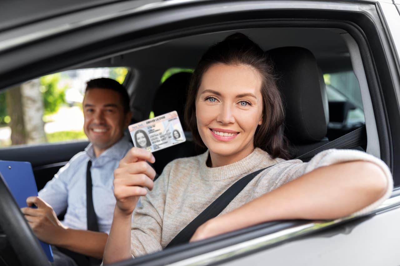 Mulher sorridente dentro de um carro. Ela está sentada no banco do motorista e mostrando a sua CNH pelo vidro, que está aberto. Ao seu lado, no banco do passageiro, há um homem, também sorridente.