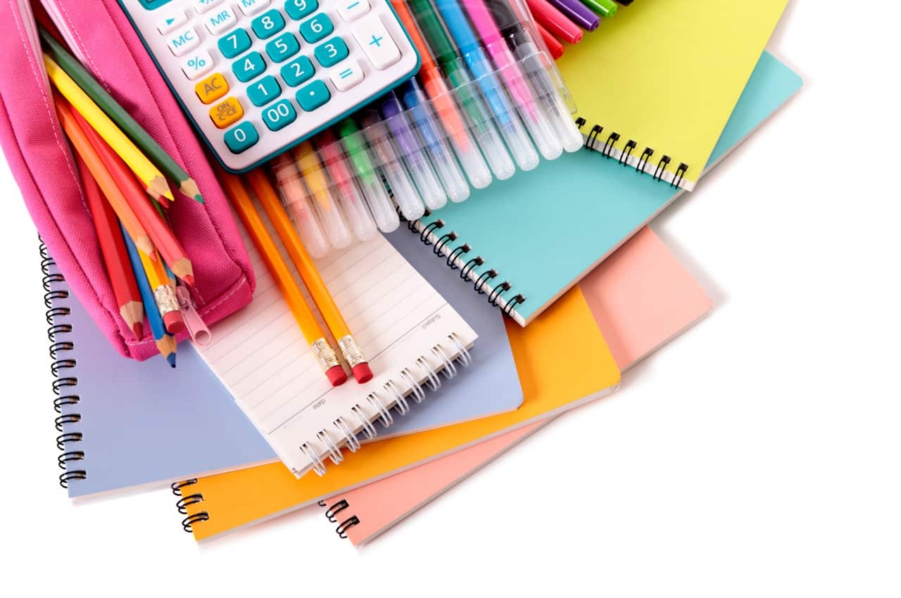 Materiais escolares, como cadernos, lápis, canetas e calculadora empilhados sobre fundo branco.