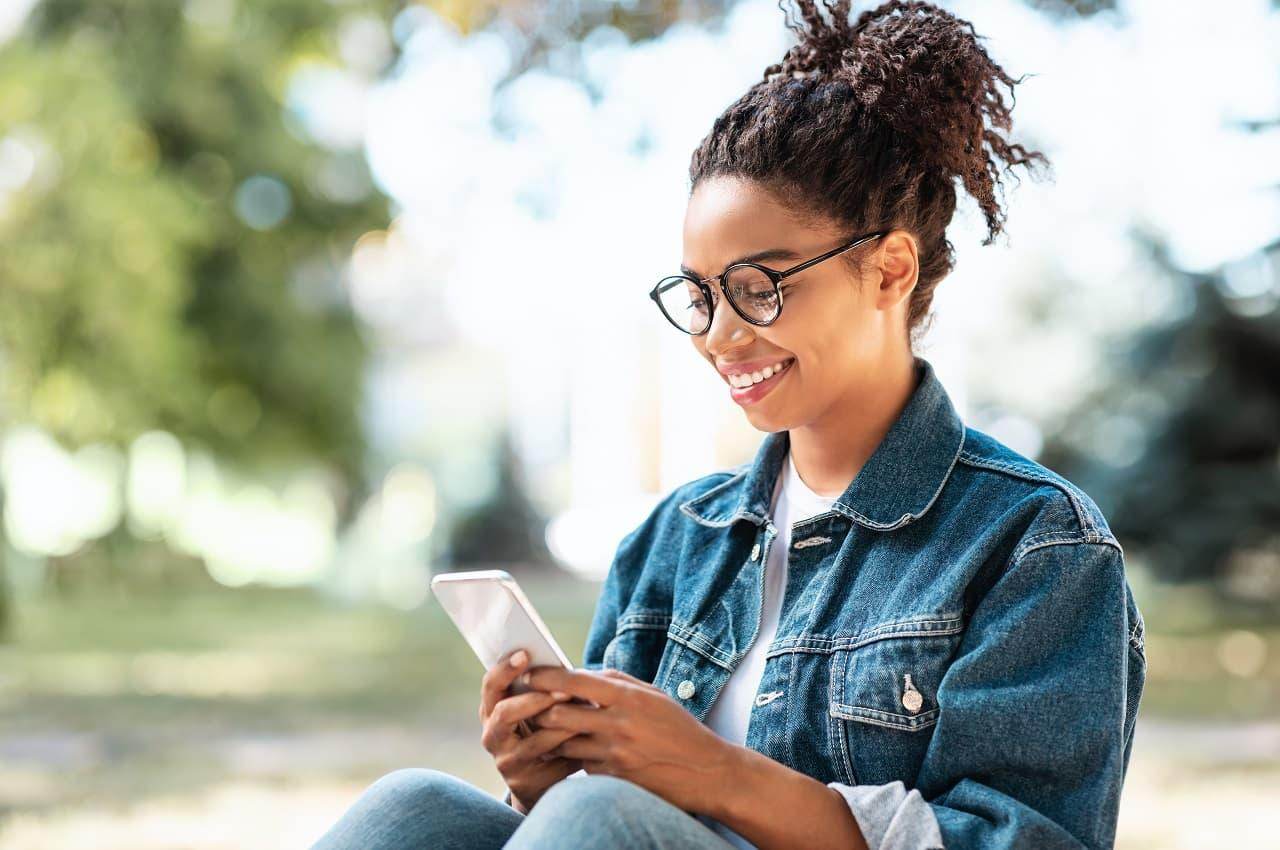 Mulher sorridente sentada em um parque e pesquisando no celular como funciona empréstimo consignado clt. Ela veste jaqueta e calça jeans, com uma blusa básica branca e óculos de grau.