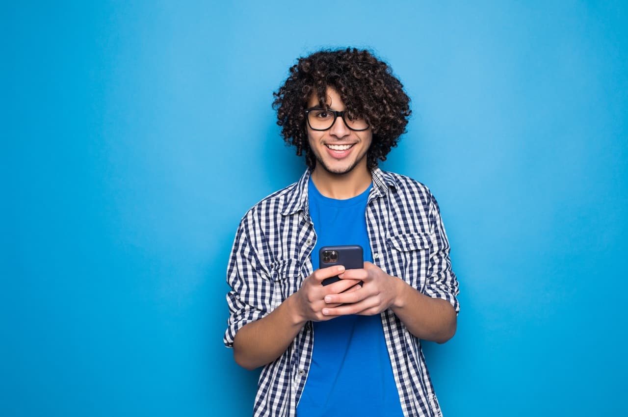 Homem jovem de óculos e cabelo cacheado, com um fundo azul, sorrindo enquanto segura um smartphone com as duas mãos. Ele está pesquisando como simular financiamento de energia solar.